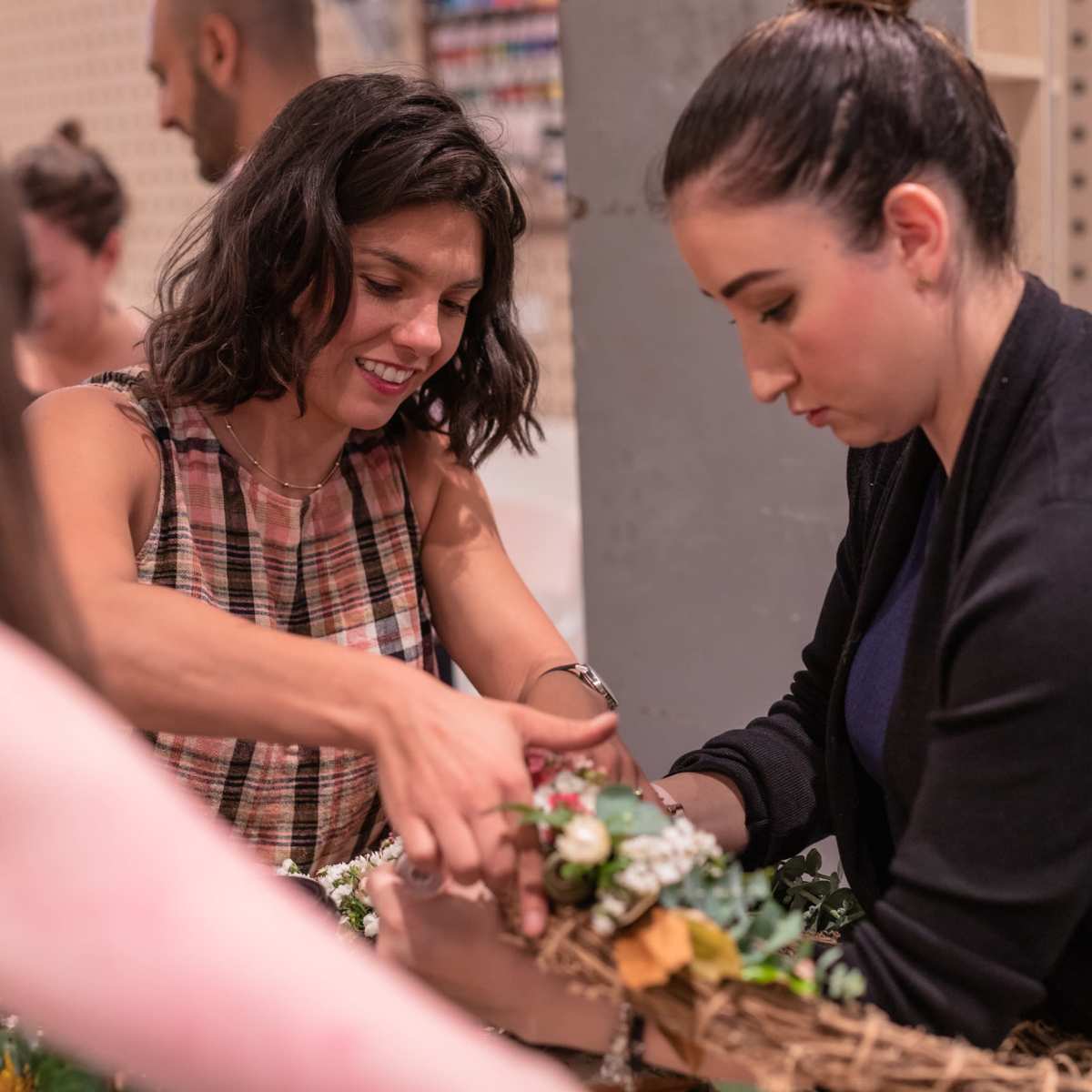 Two women collaborate on arranging flowers and greenery in a basket during a crafting session.