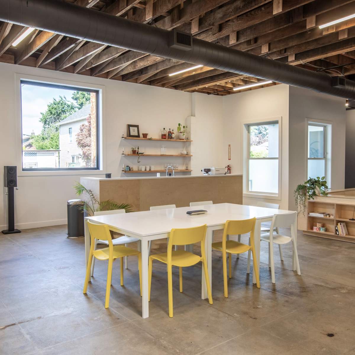 A spacious, modern interior with a white dining table surrounded by yellow and white chairs, hardwood beams overhead, and a corner featuring a low bookshelf and seating area.