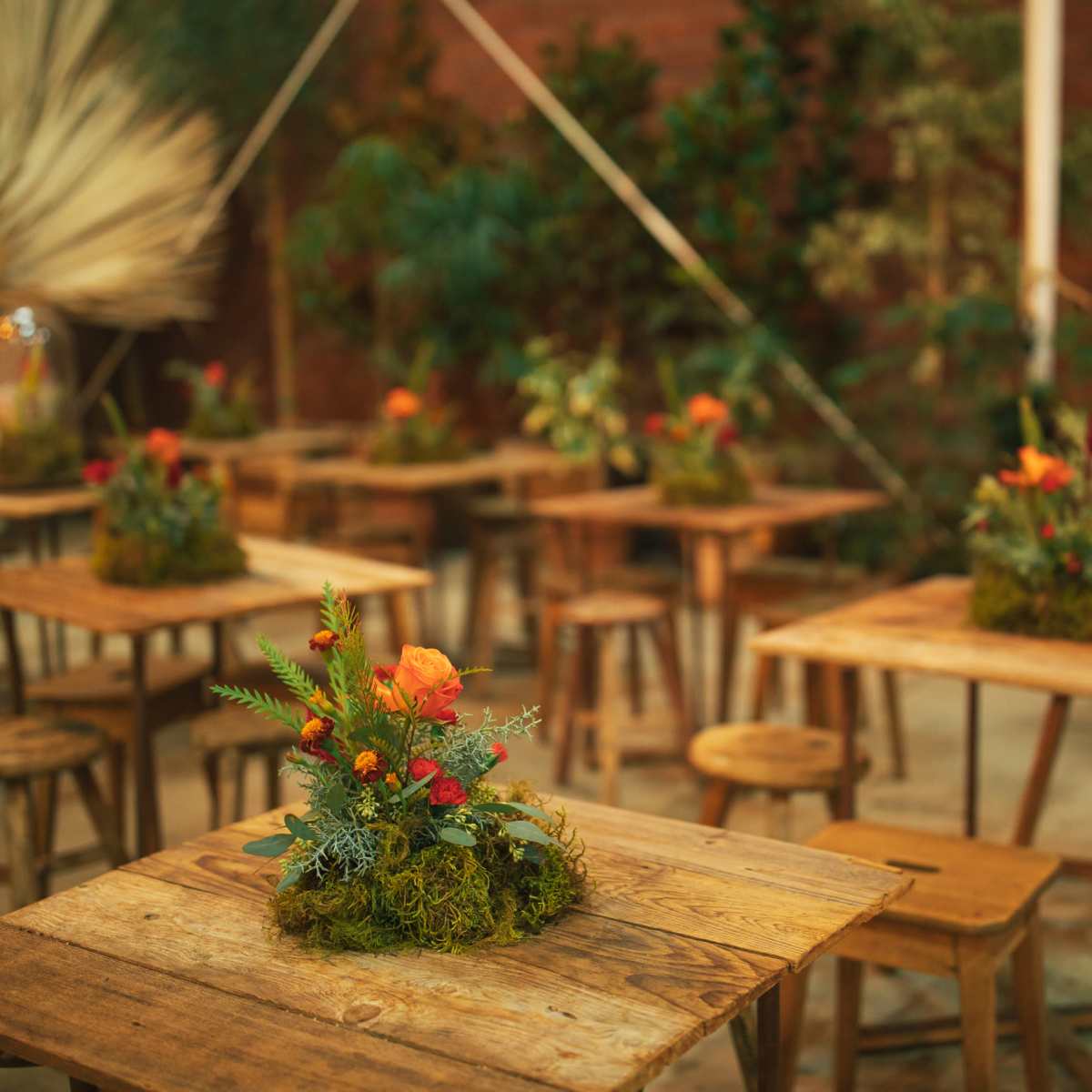 The scene features wooden tables adorned with floral arrangements in moss, surrounded by stools and a black piano in a decorated indoor space.