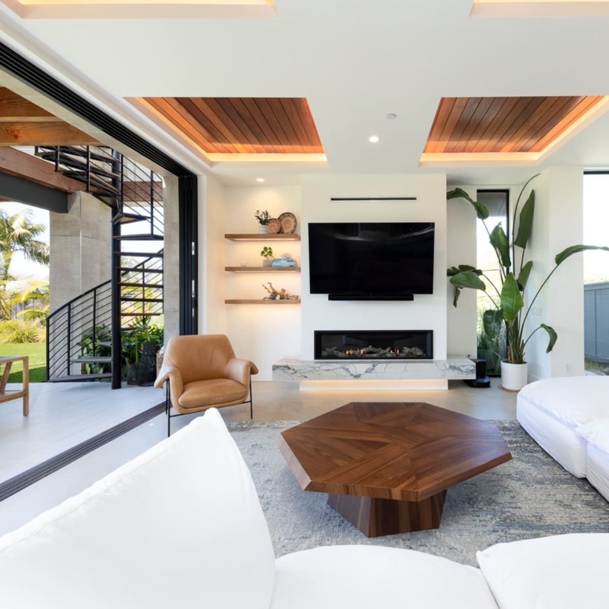 A modern living room with a white sectional sofa, a wooden coffee table, a wall-mounted television, and large windows overlooking a patio with greenery.