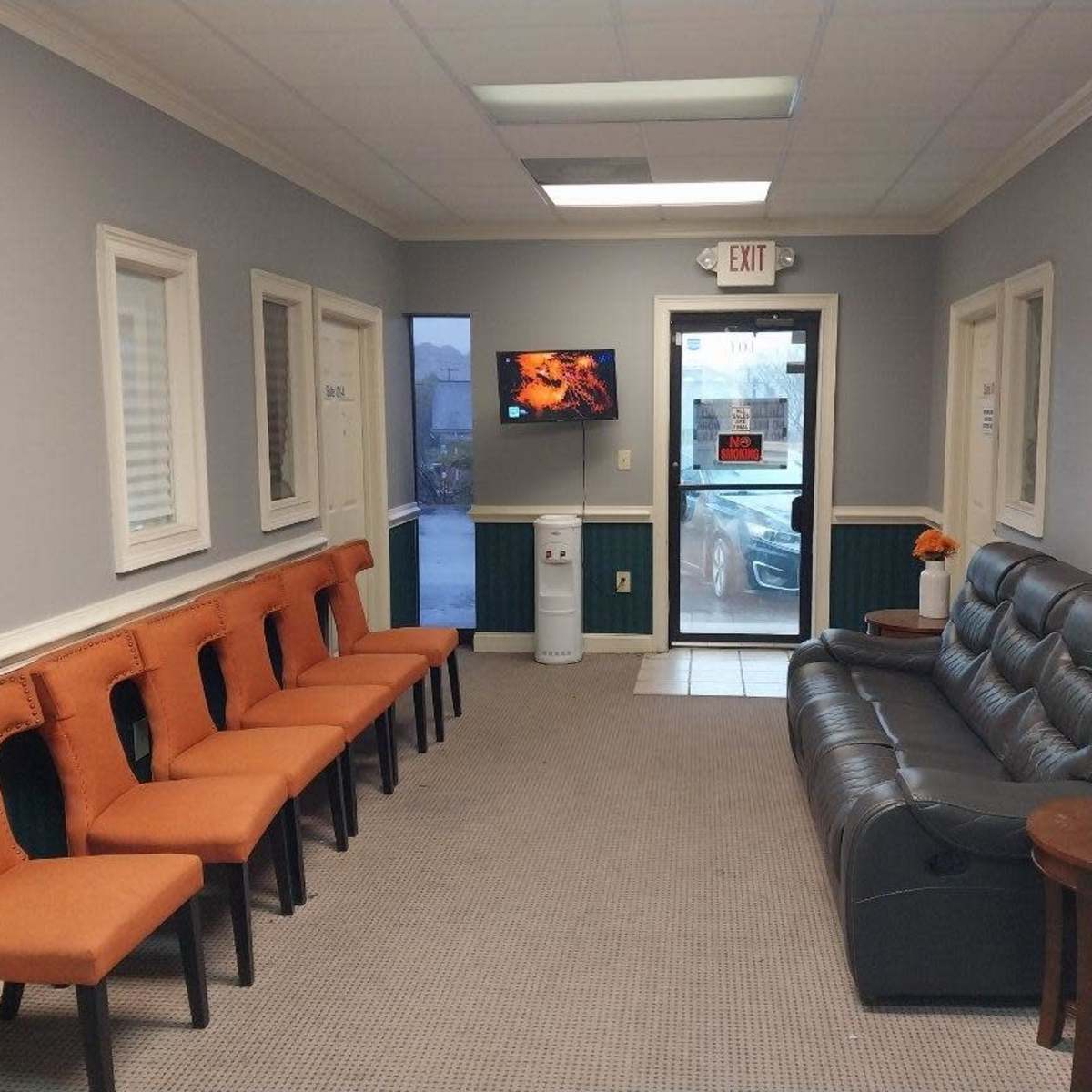 A waiting room with several orange chairs along one wall, a black sofa, and a TV mounted on the opposite wall near a glass door.