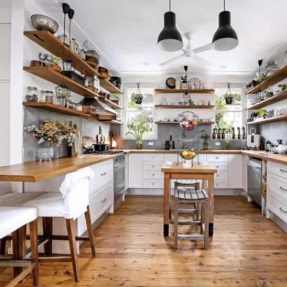 The kitchen features white cabinets, wooden shelves with various plants and decor, and a central table with chairs, all set on a wooden floor.