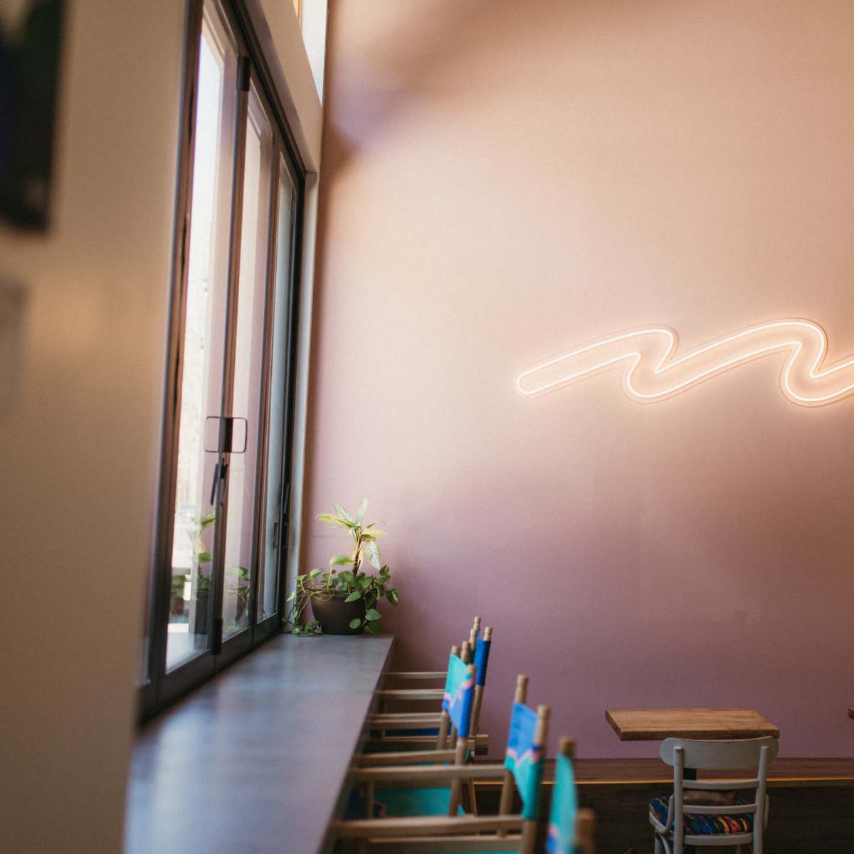 A café interior features a pink wall with a wavy neon light above a row of tables and chairs.
