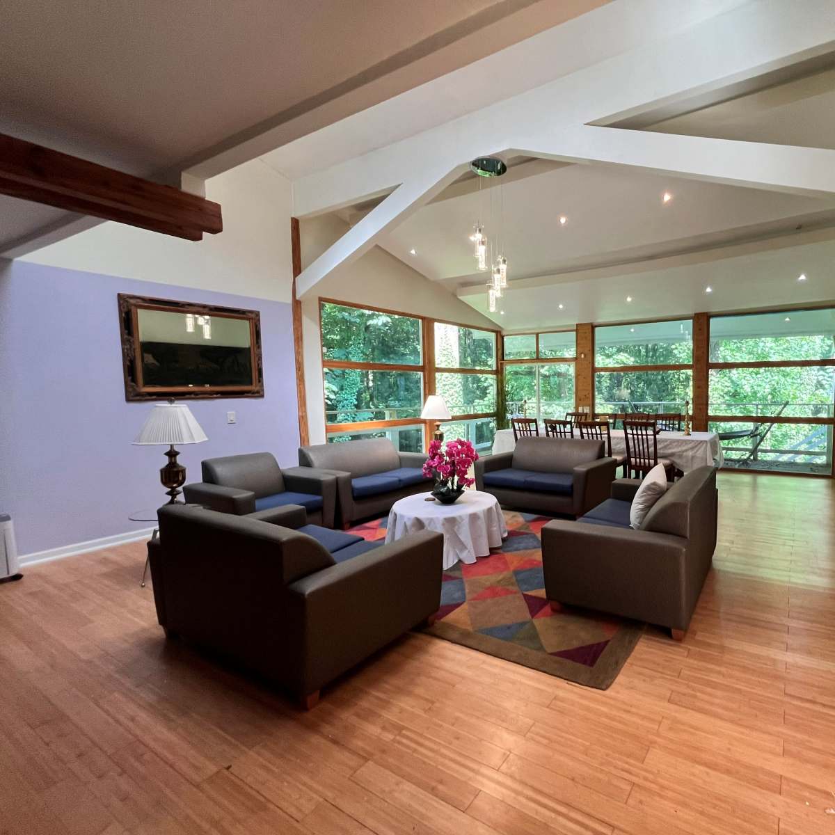 A spacious living area with large windows overlooking greenery, featuring modern sofas arranged around a coffee table and a dining area in the background.