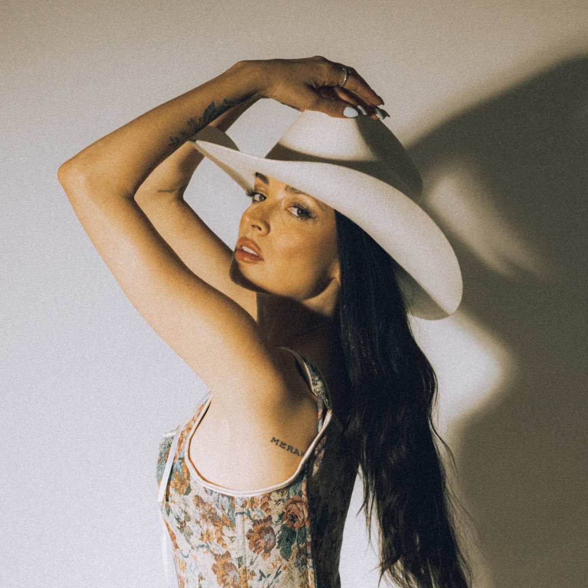 A woman with long black hair poses sideways while wearing a floral crop top and a wide-brimmed white hat.
