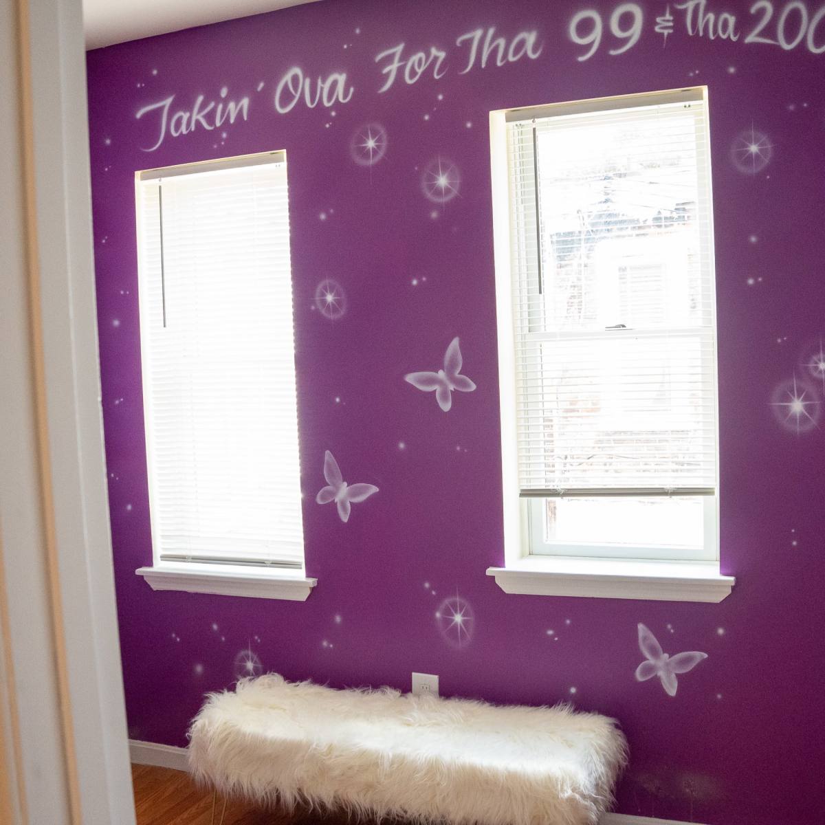A small room with purple walls decorated with white butterflies and text, featuring two windows and a white fur bench.