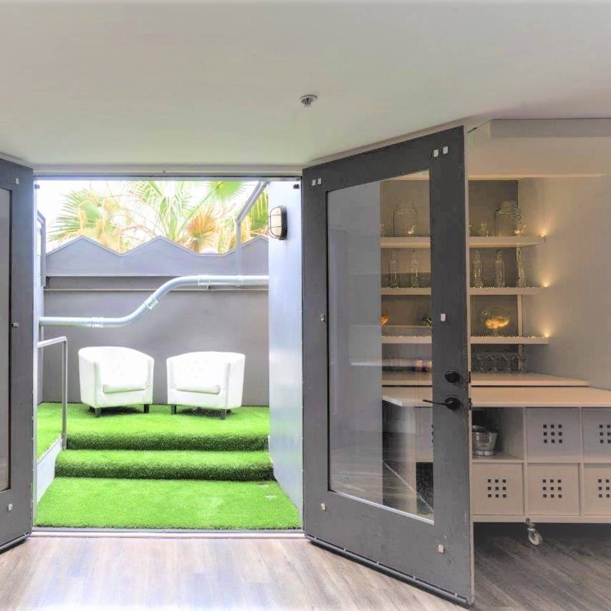 A room with double doors leading to a small outdoor area featuring two white chairs on green artificial grass.