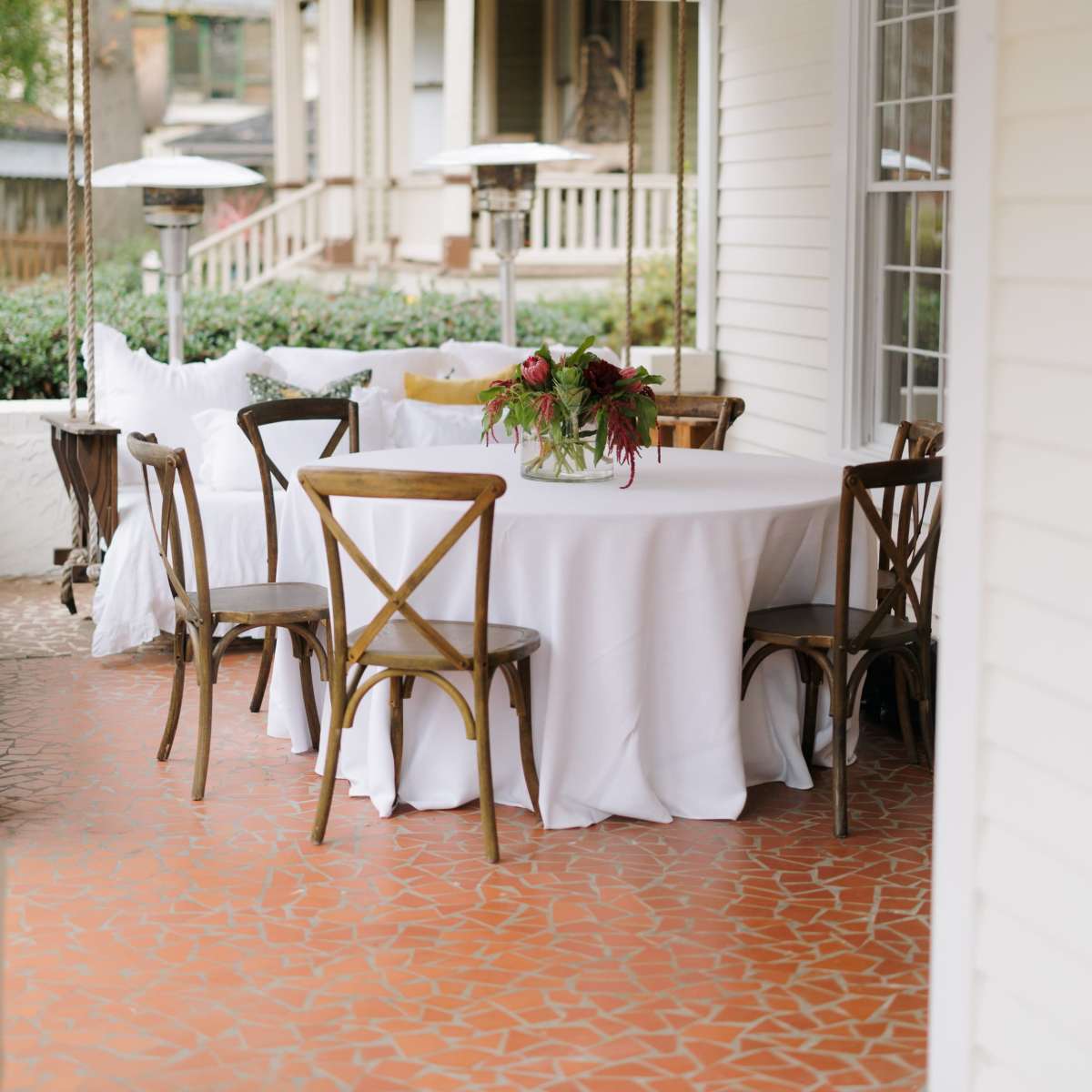 A round table with a white tablecloth is set in a porch area surrounded by six wooden chairs, with a vase of flowers at the center.