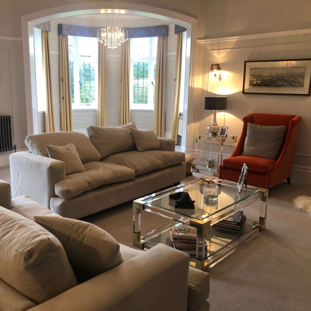 The living room features two beige sofas, an orange armchair, a glass coffee table, and large windows with sheer curtains.