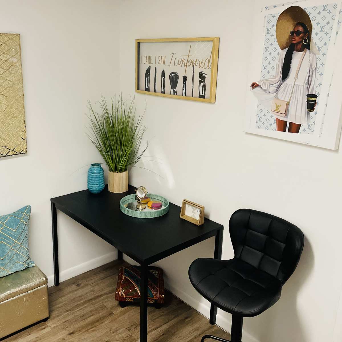 A small, stylish workspace featuring a black desk, a chair, decorative pillows, and framed artwork on the walls.