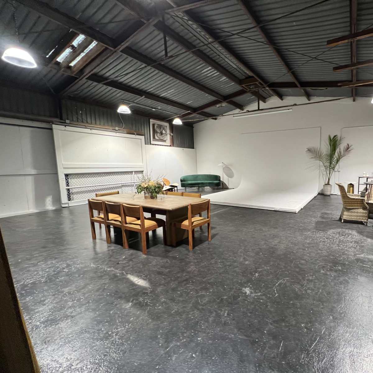 A spacious industrial-style room with a large empty backdrop, a dining table, a green couch, and various seating arrangements.