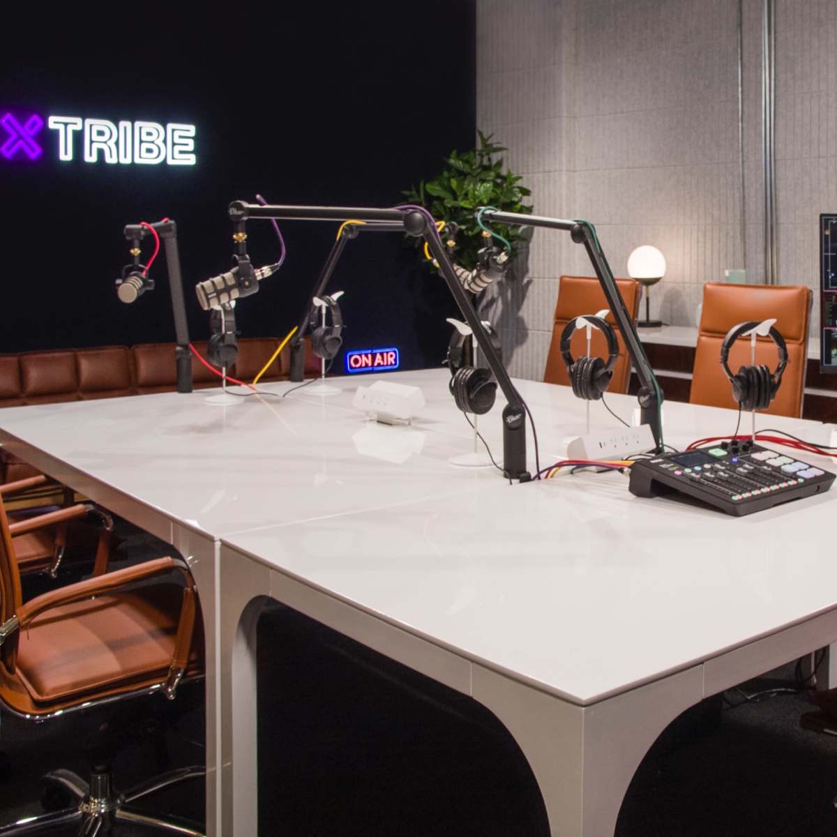 A modern podcasting studio with a white table, eight brown leather chairs, audio equipment, and multiple monitors displaying video feeds.