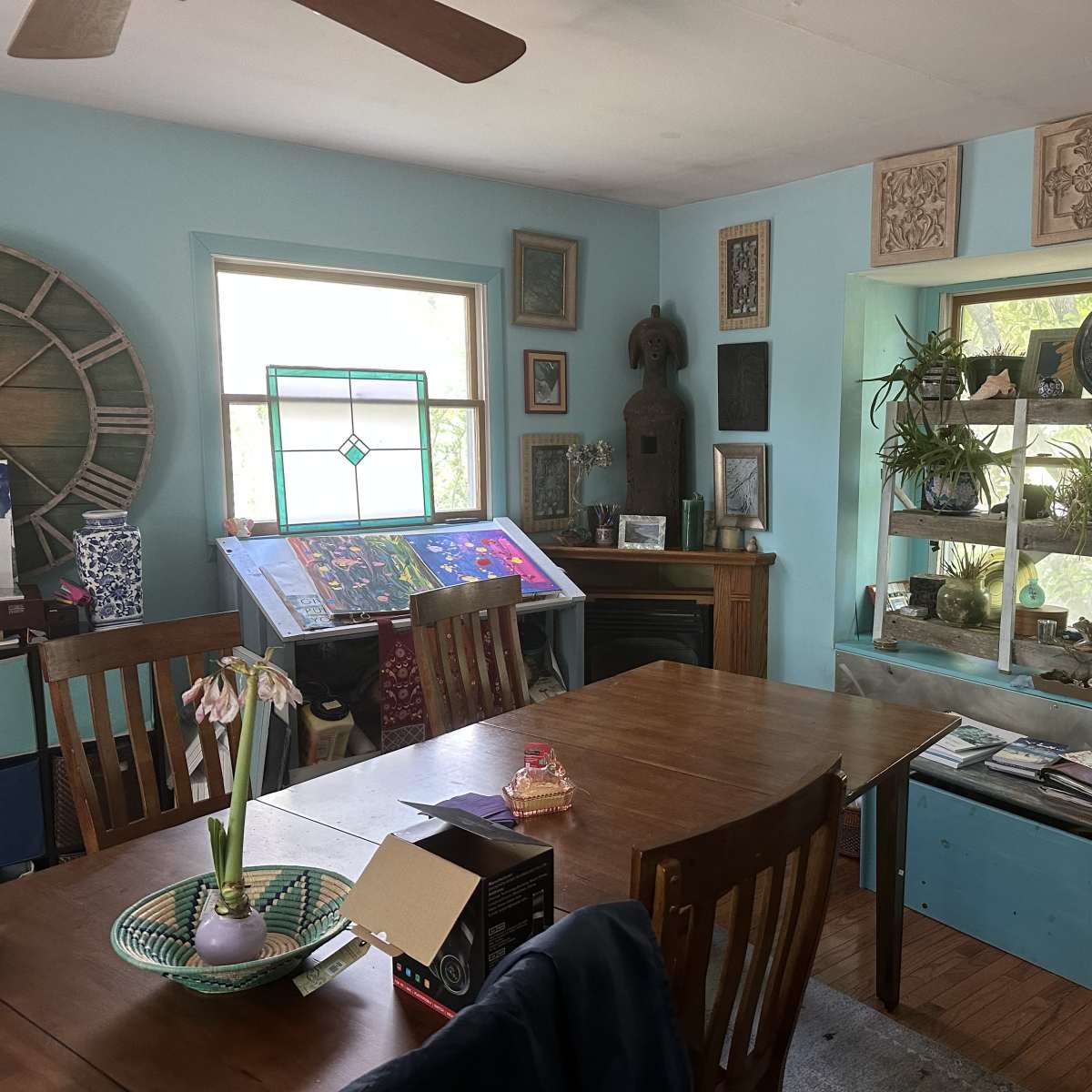 The scene features a brightly lit dining area with a wooden table surrounded by chairs, a fireplace, and shelves displaying various plants and decorative objects.