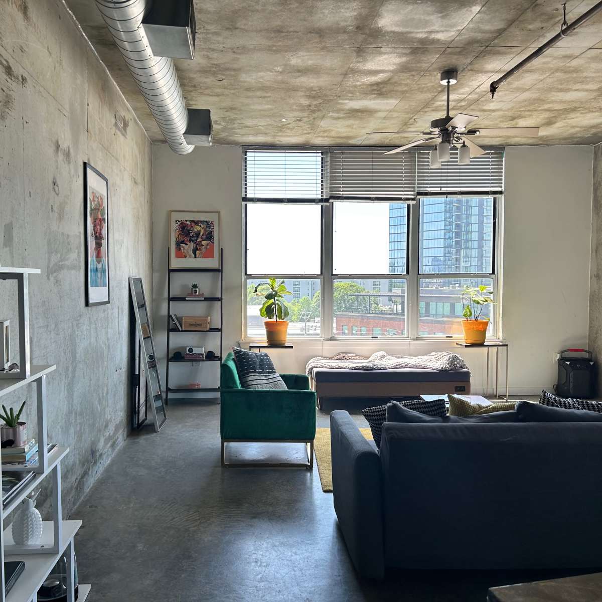 A modern apartment with concrete walls, large windows, and a green sofa facing a well-lit area with a bed and indoor plants.