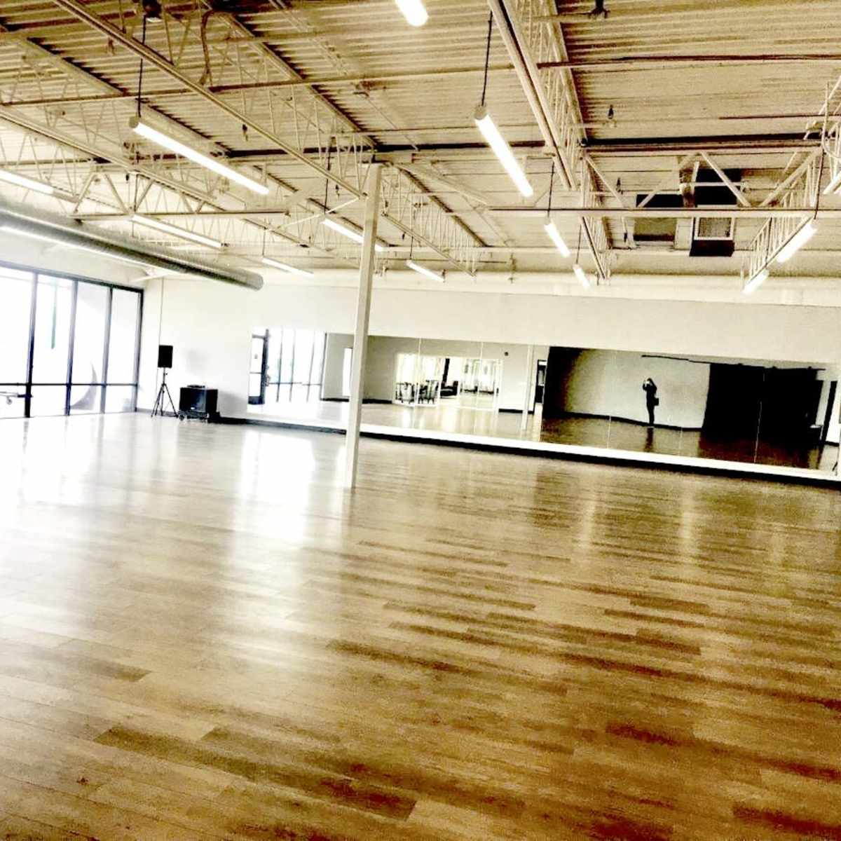 An empty dance studio with large windows, a mirrored wall, and wooden flooring.