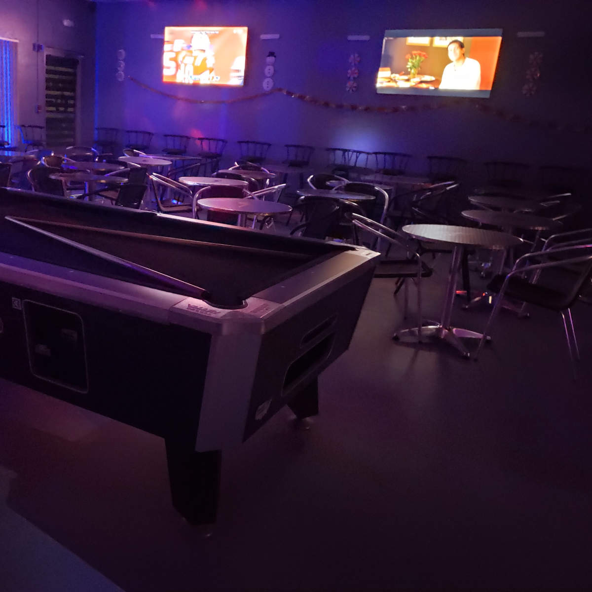 A pool table sits in a dimly lit room with rows of empty tables and chairs, and a television displaying a show in the background.