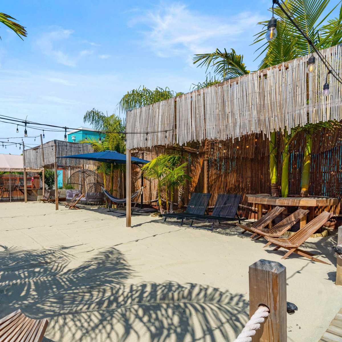 A sand-covered outdoor area with several hammocks, palm trees, and wooden seating arranged under a canopy structure.