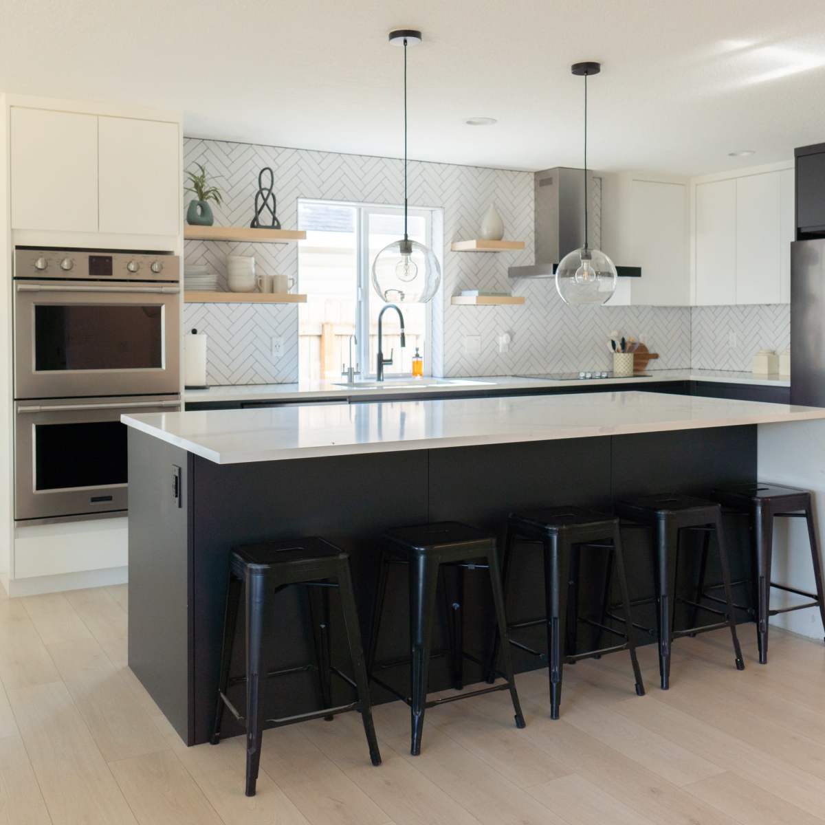 A modern kitchen features a large island with black stools, stainless steel appliances, and light wood flooring.