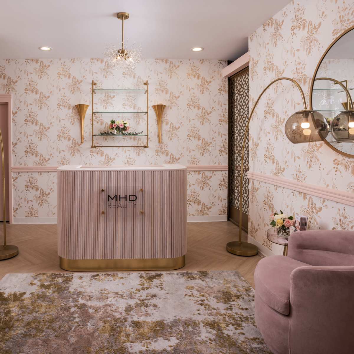 A modern beauty salon reception area with a curved front desk, pink chairs, decorative mirrors, and floral wallpaper.