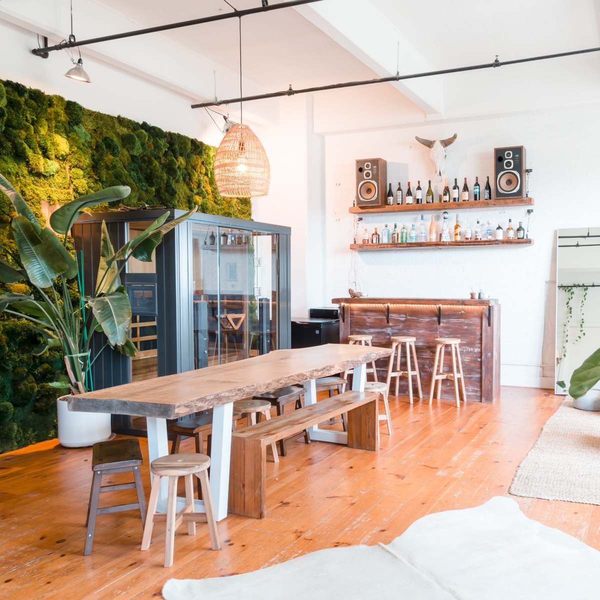 A spacious, modern living area featuring a large wooden dining table, a bar with liquor bottles, a green moss wall, and various plants.