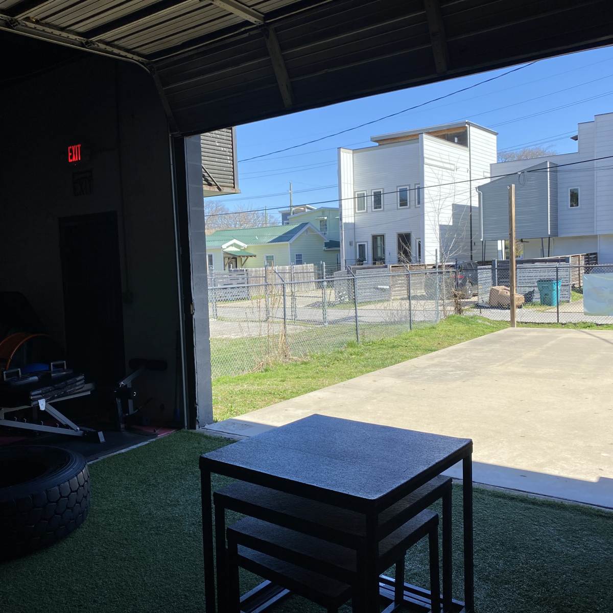 An open garage space with gym equipment on one side and a view of a fenced outdoor area and buildings in the background.