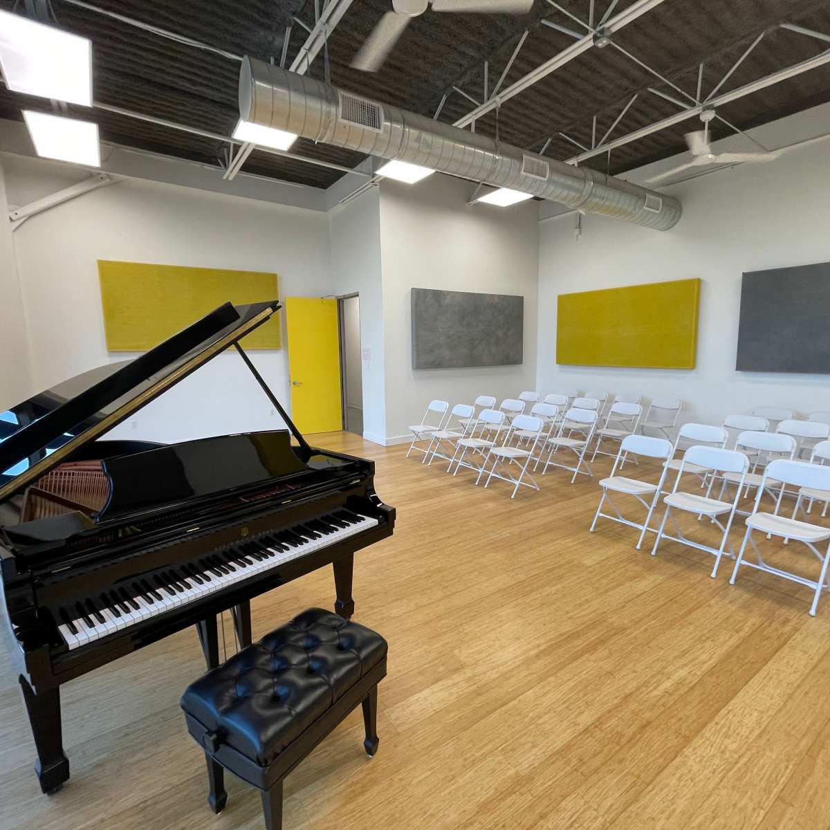 A black grand piano with a matching bench is positioned next to rows of white folding chairs in a well-lit room with yellow and gray wall panels.
