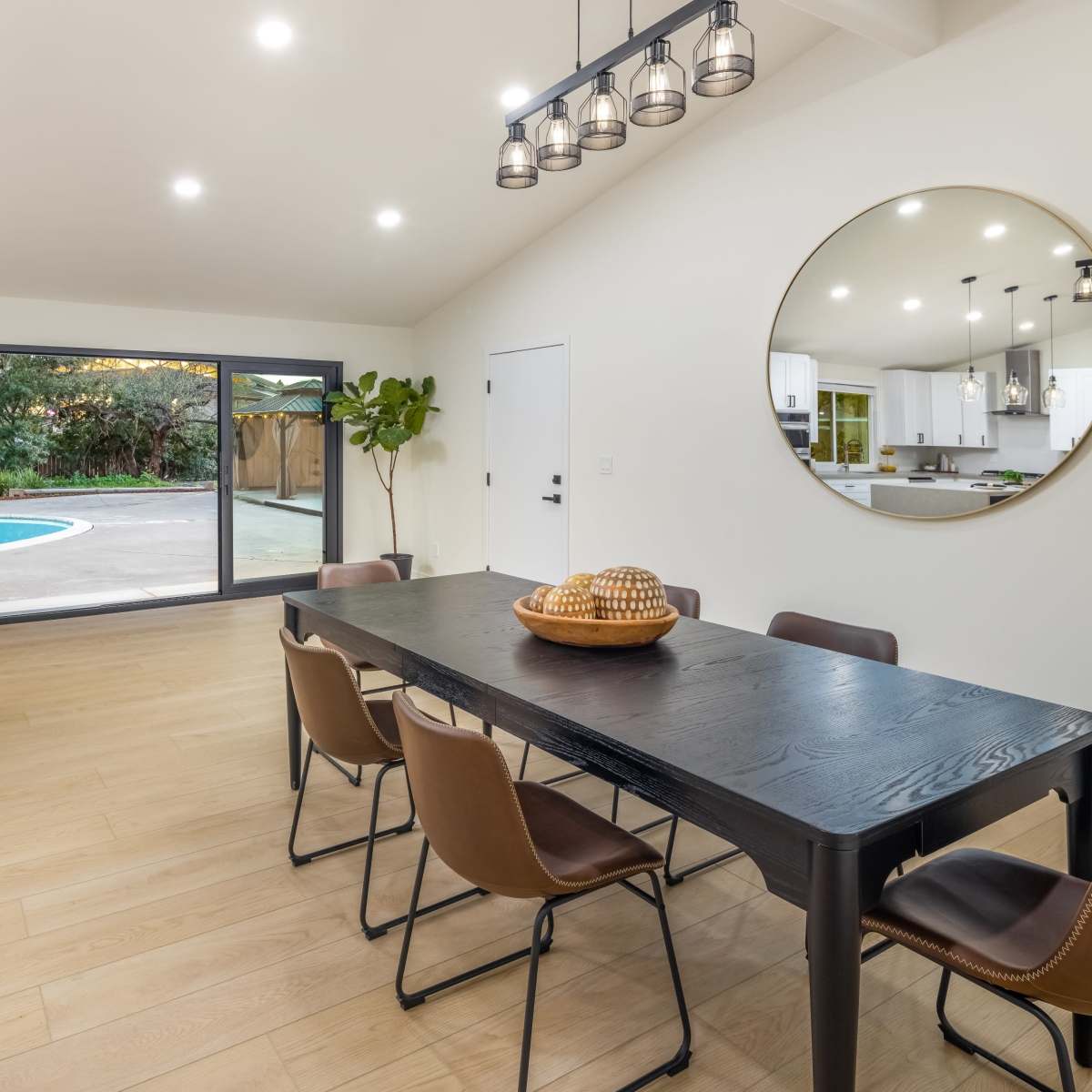 A dining area features a black wooden table surrounded by brown chairs, with large windows providing a view of a backyard pool.