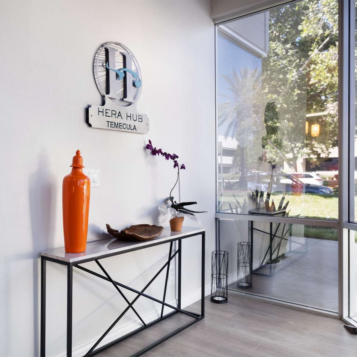 A bright entryway with a decorative table displaying an orange vase, an orchid plant, and a sign for Hera Hub Temecula on the wall.