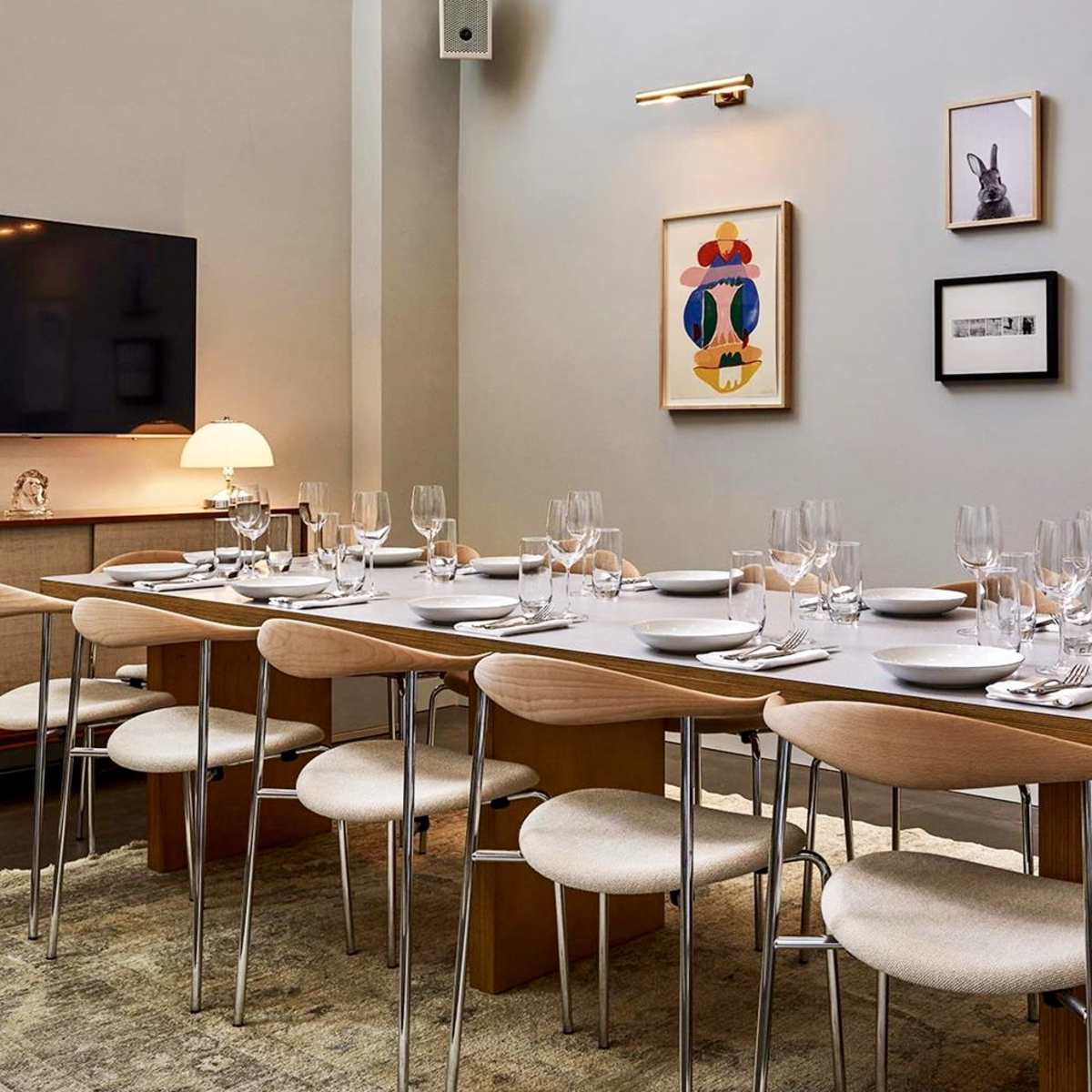 A modern dining room features a long table set for dinner, surrounded by chairs and decorated with framed artwork on the walls.