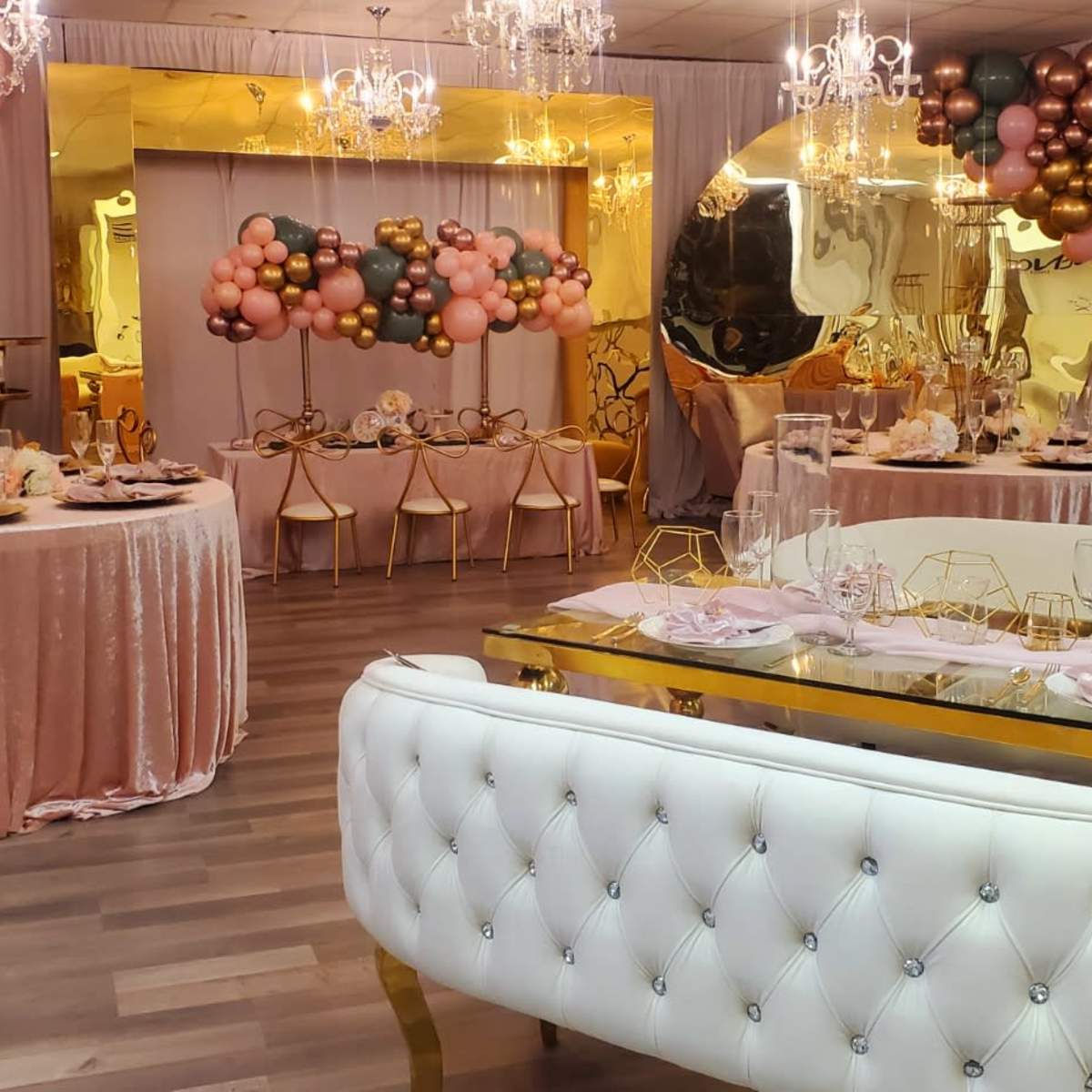 An elegantly decorated event space featuring tables adorned with pink and gold accents, surrounded by balloon arrangements and chandeliers.
