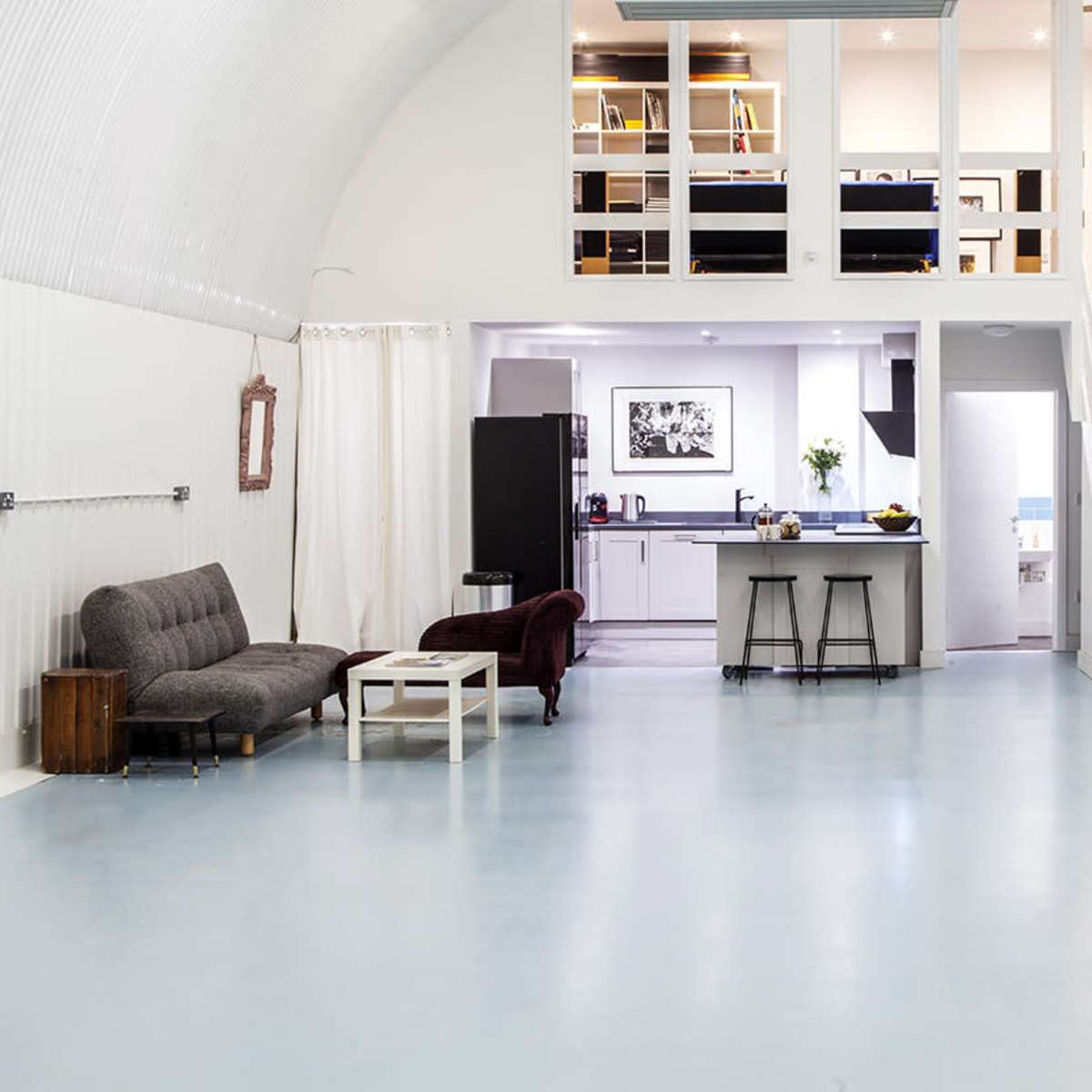 A spacious, modern room with a high ceiling, featuring a living area with a gray sofa, a burgundy chair, and a small white table, leading to a kitchenette and a staircase.