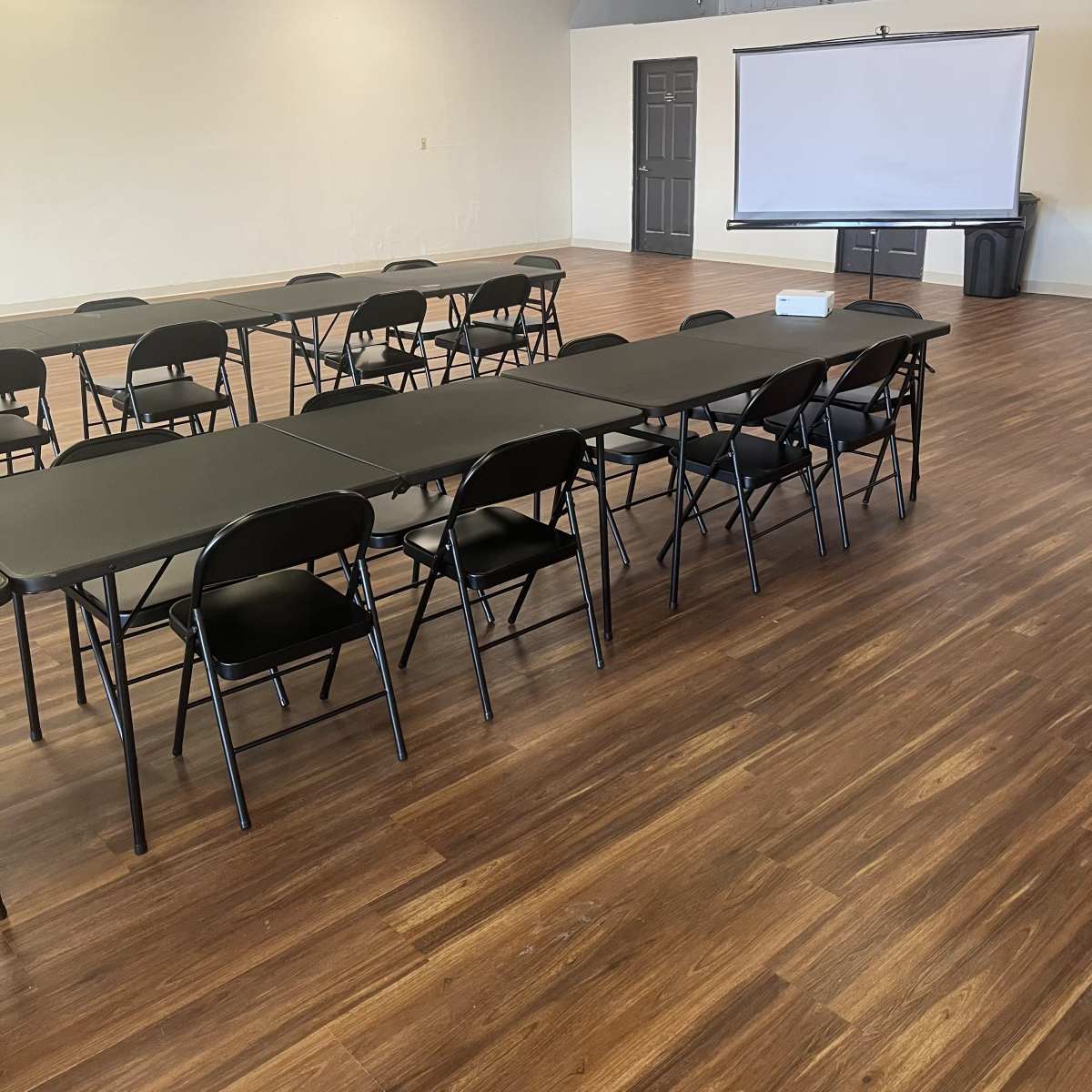 A spacious room with black rectangular tables and folding chairs arranged in rows, along with a blank projection screen mounted on a wall.