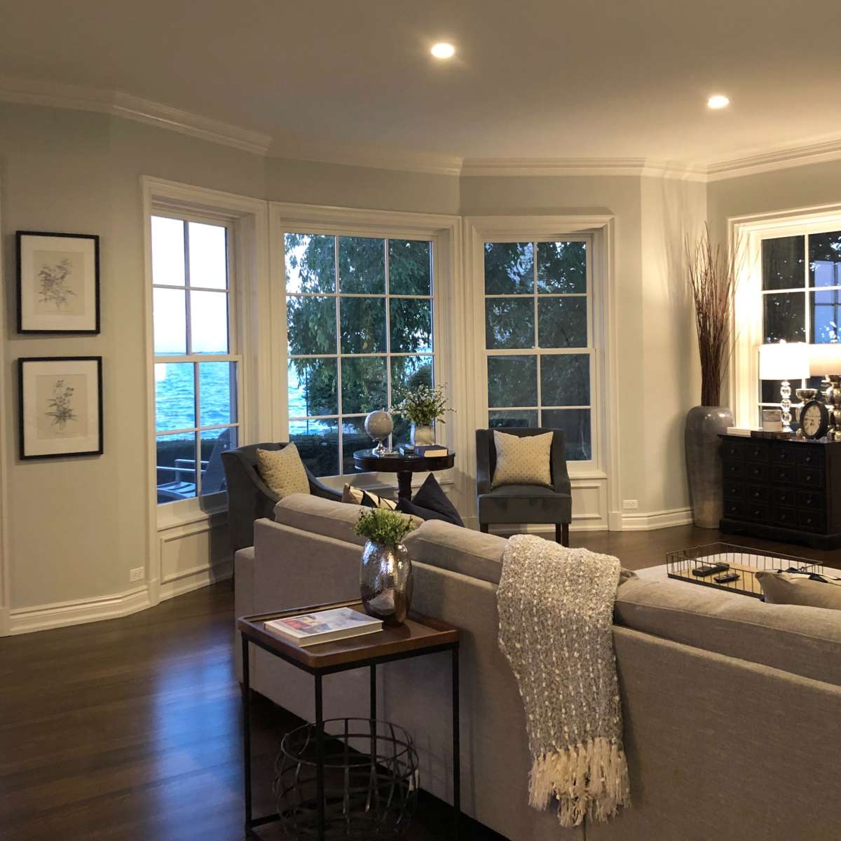 The living room features large windows overlooking a body of water, with a sofa, decorative pillows, a coffee table, and a media console.