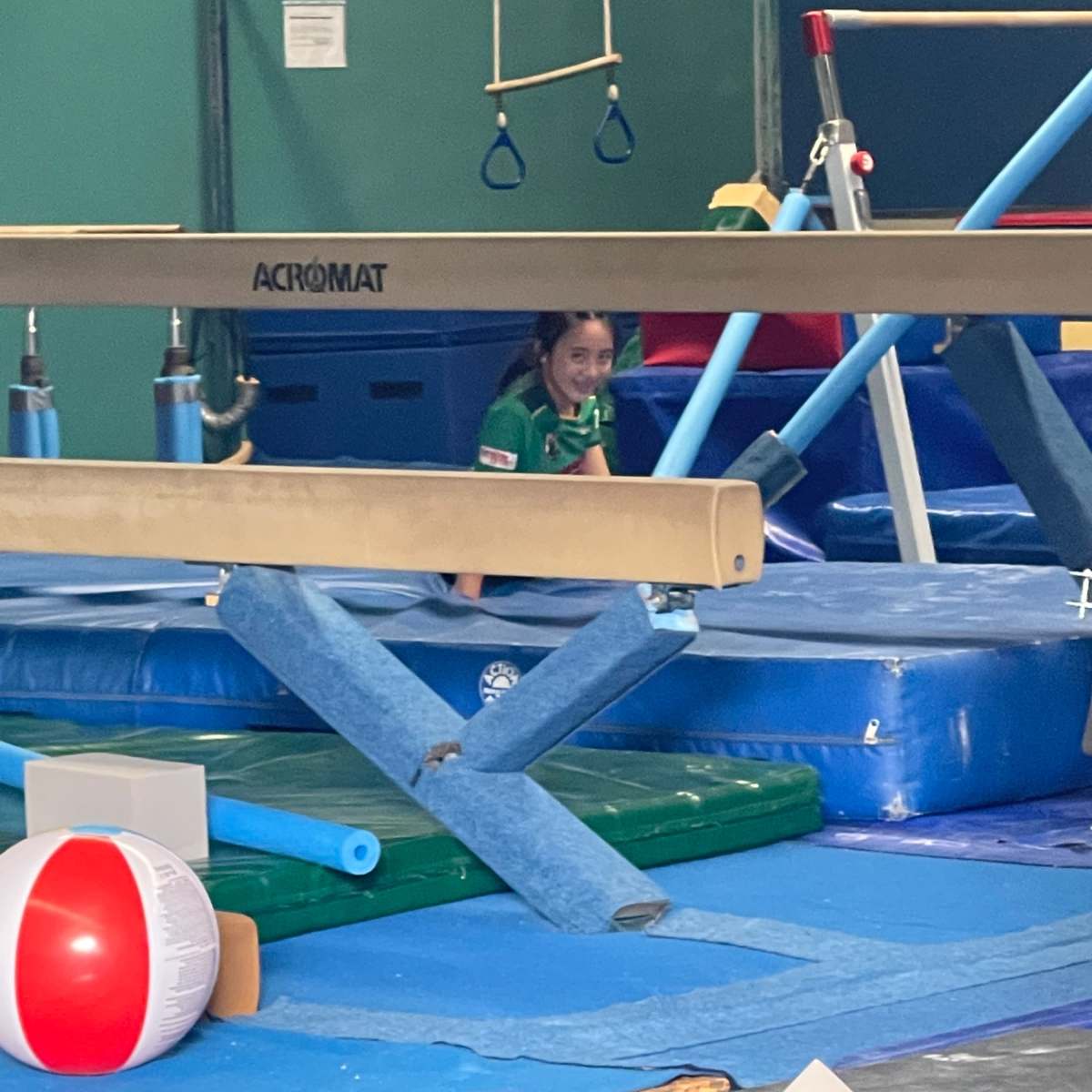A young gymnast is practicing in a gymnasium, positioned near a balance beam and various colorful gymnastic equipment.