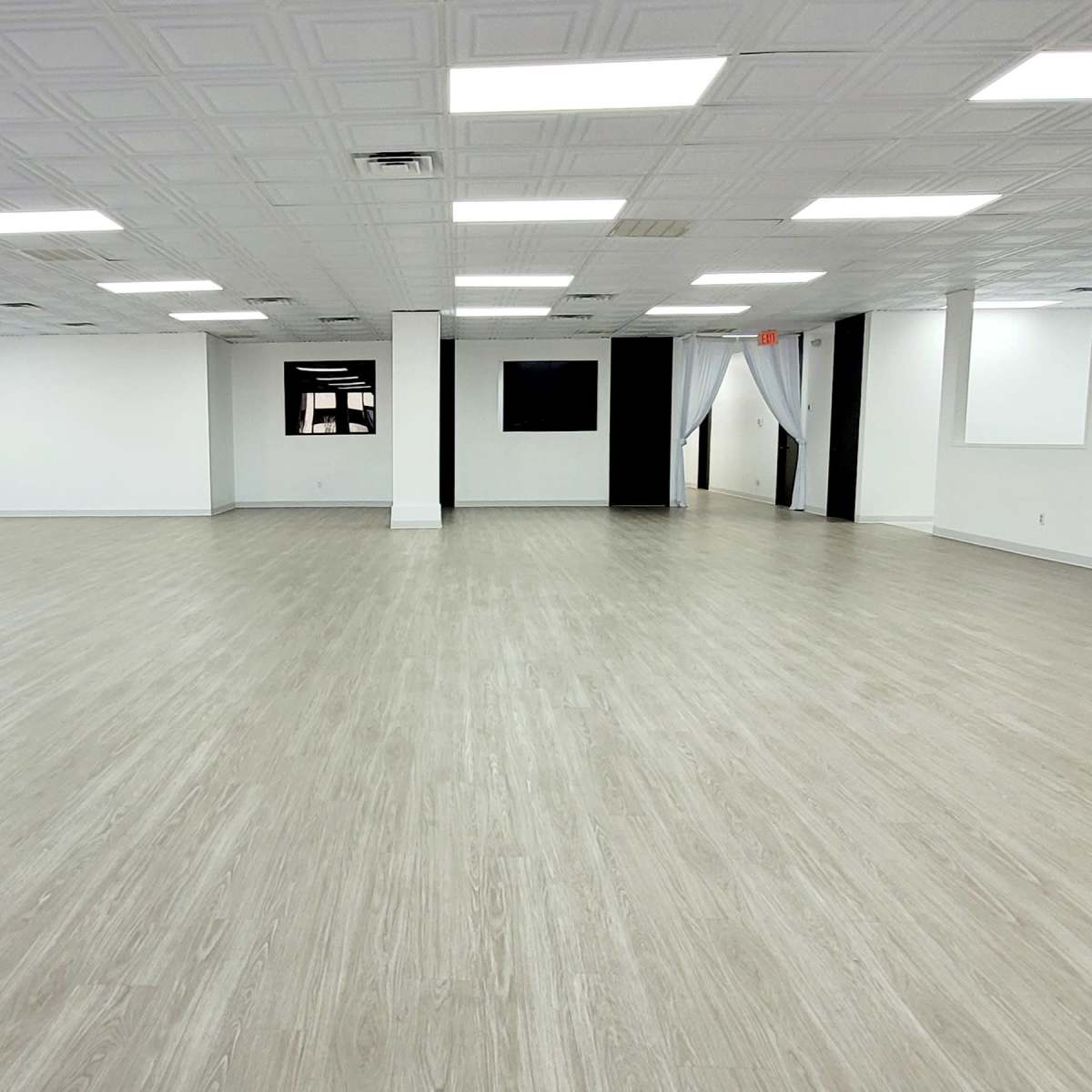 A spacious, empty room with light-colored, wooden flooring and white walls, illuminated by overhead lighting.