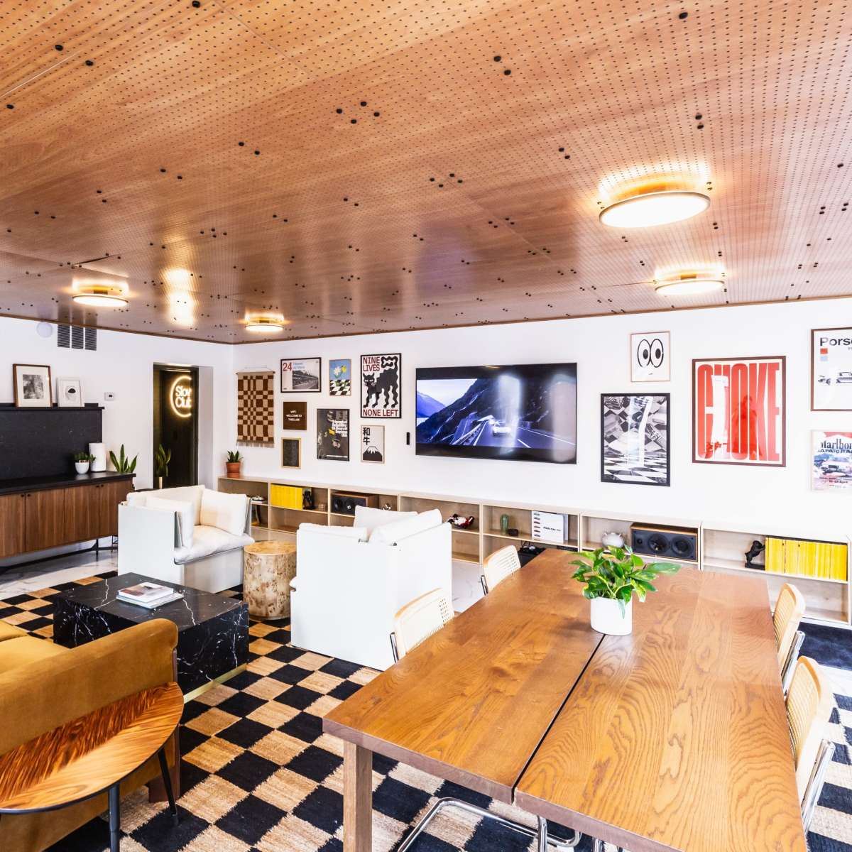 A modern lounge area with a wooden ceiling, a black and white checkered floor, comfortable seating, a large television, and various artwork on the walls.