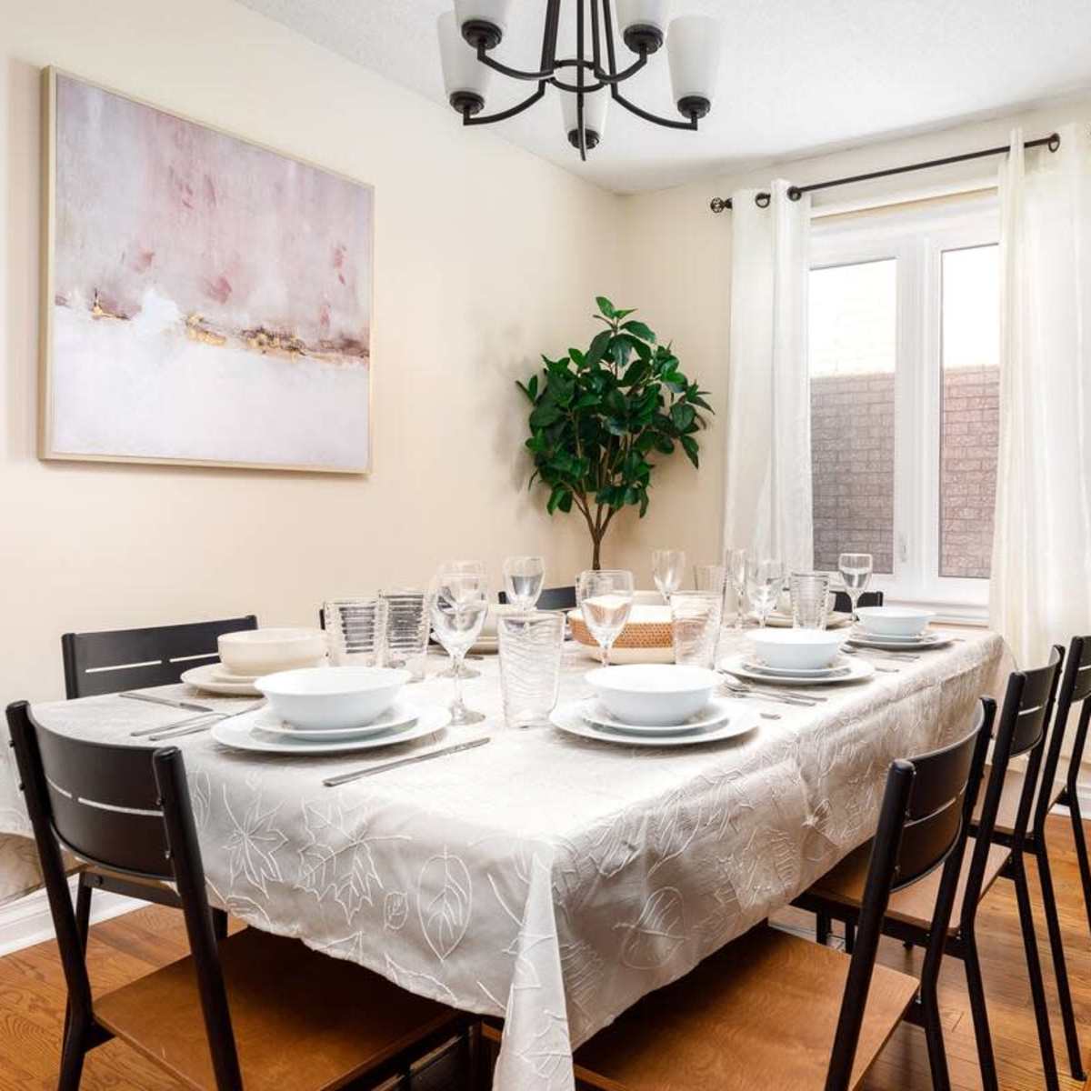 A dining table is set for a meal, featuring plates, glasses, and cutlery, illuminated by a chandelier in a light-colored room.
