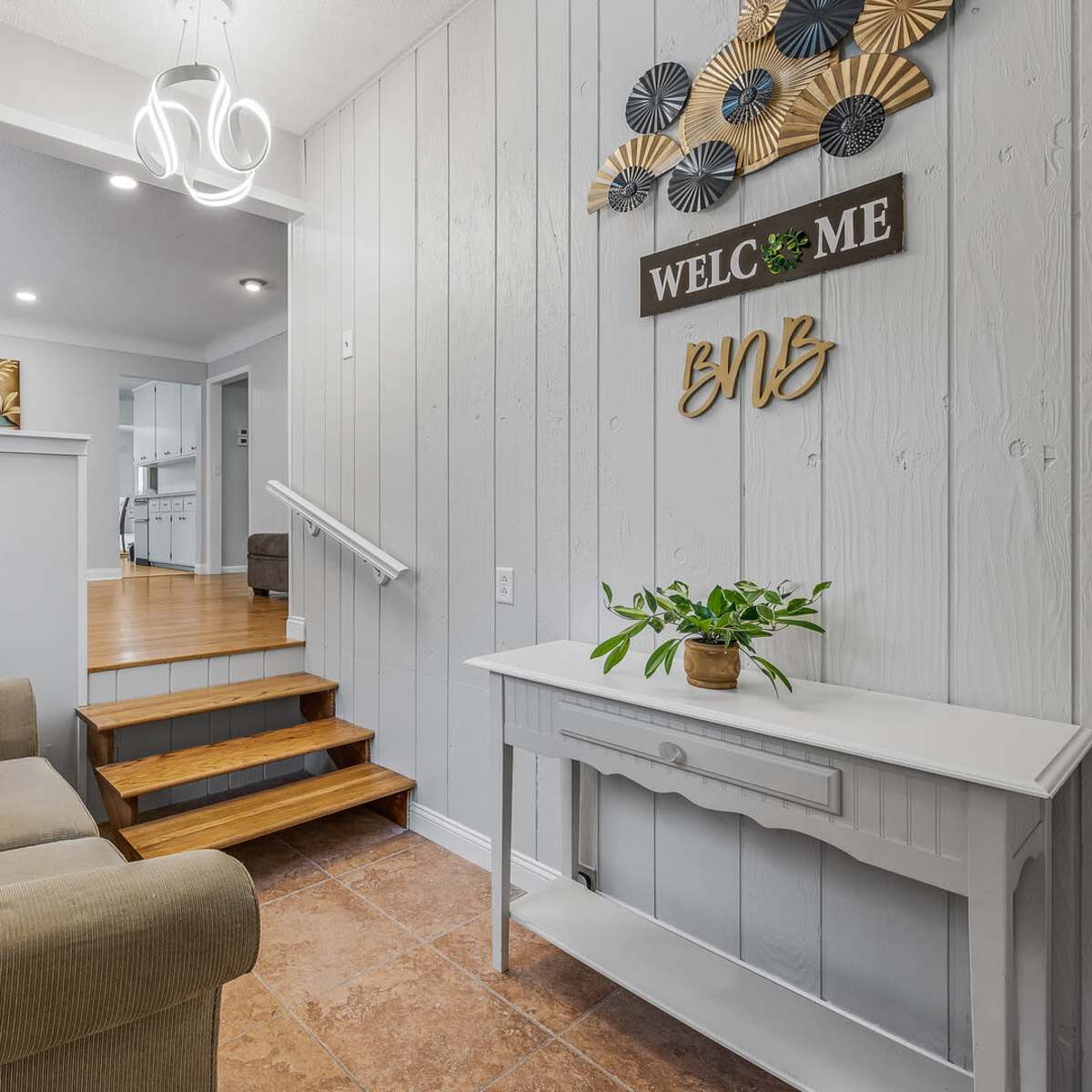 A small entryway with a beige couch, wooden stairs leading up, and a decorative wall featuring a welcome sign and potted plants.
