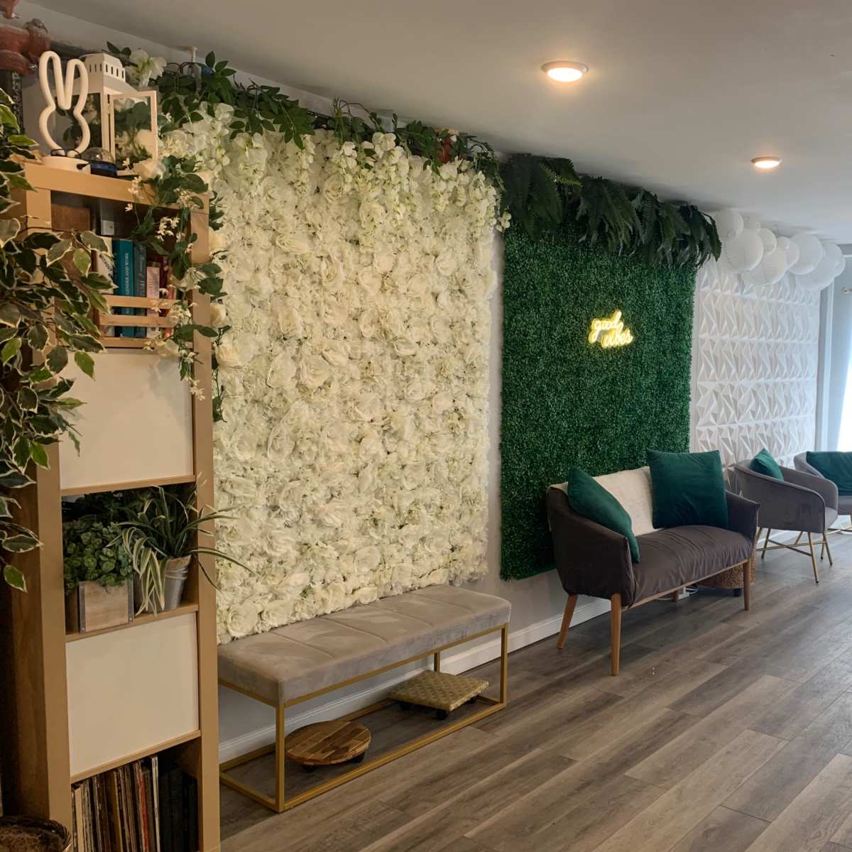 The scene features a modern indoor space with faux floral walls, greenery, and stylish seating arranged along a wood-paneled floor.