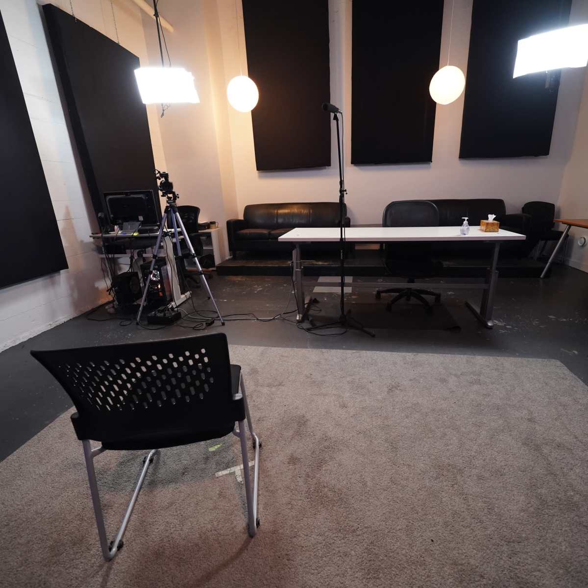 A recording studio with a microphone on a stand, a camera setup, and a single chair facing a table in a minimalistic, well-lit space.