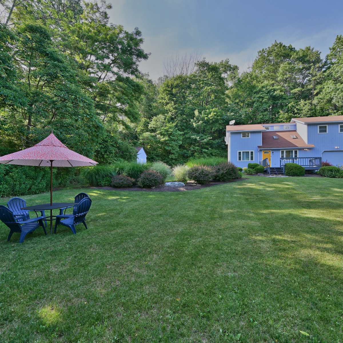 A blue two-story house is surrounded by trees, with a green lawn featuring a round table and chairs underneath a pink umbrella.