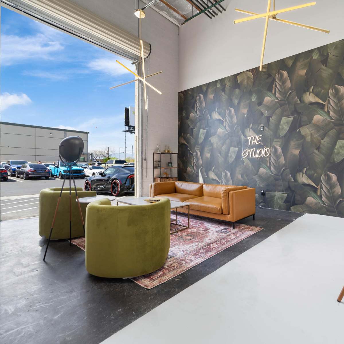 A modern studio space featuring a leather couch, two green chairs, a large plant mural on one wall, and large windows allowing natural light to enter.