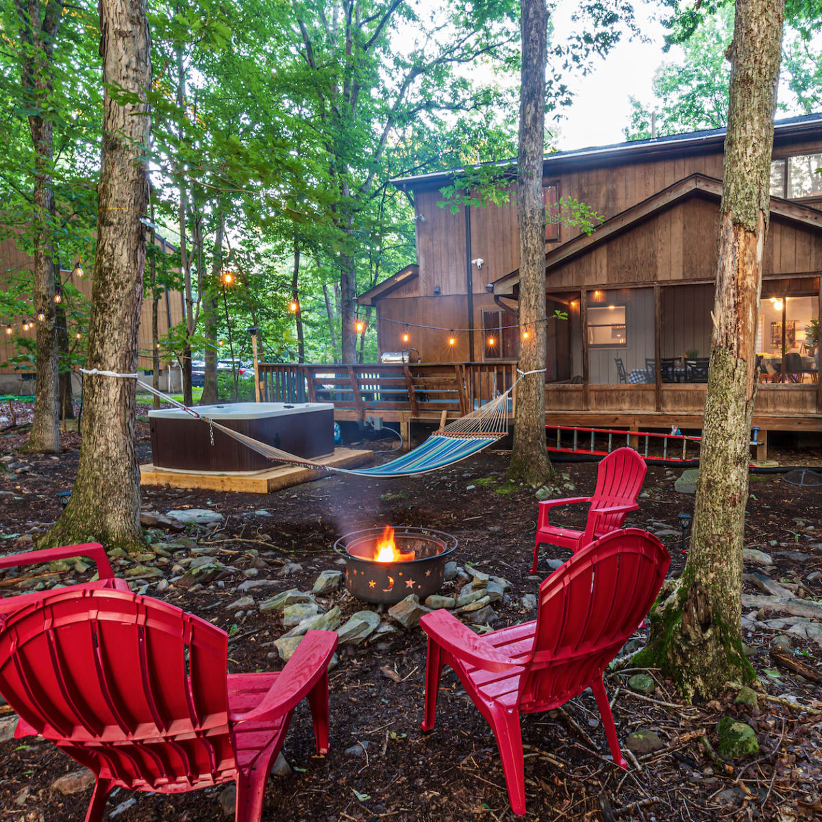 A cozy cabin surrounded by trees features a fire pit with red chairs, a hammock nearby, and a deck with outdoor seating visible.