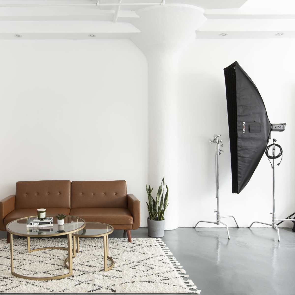 A modern studio living space with a brown couch, two circular tables, a plant, and a backdrop of colored paper rolls against a white wall.