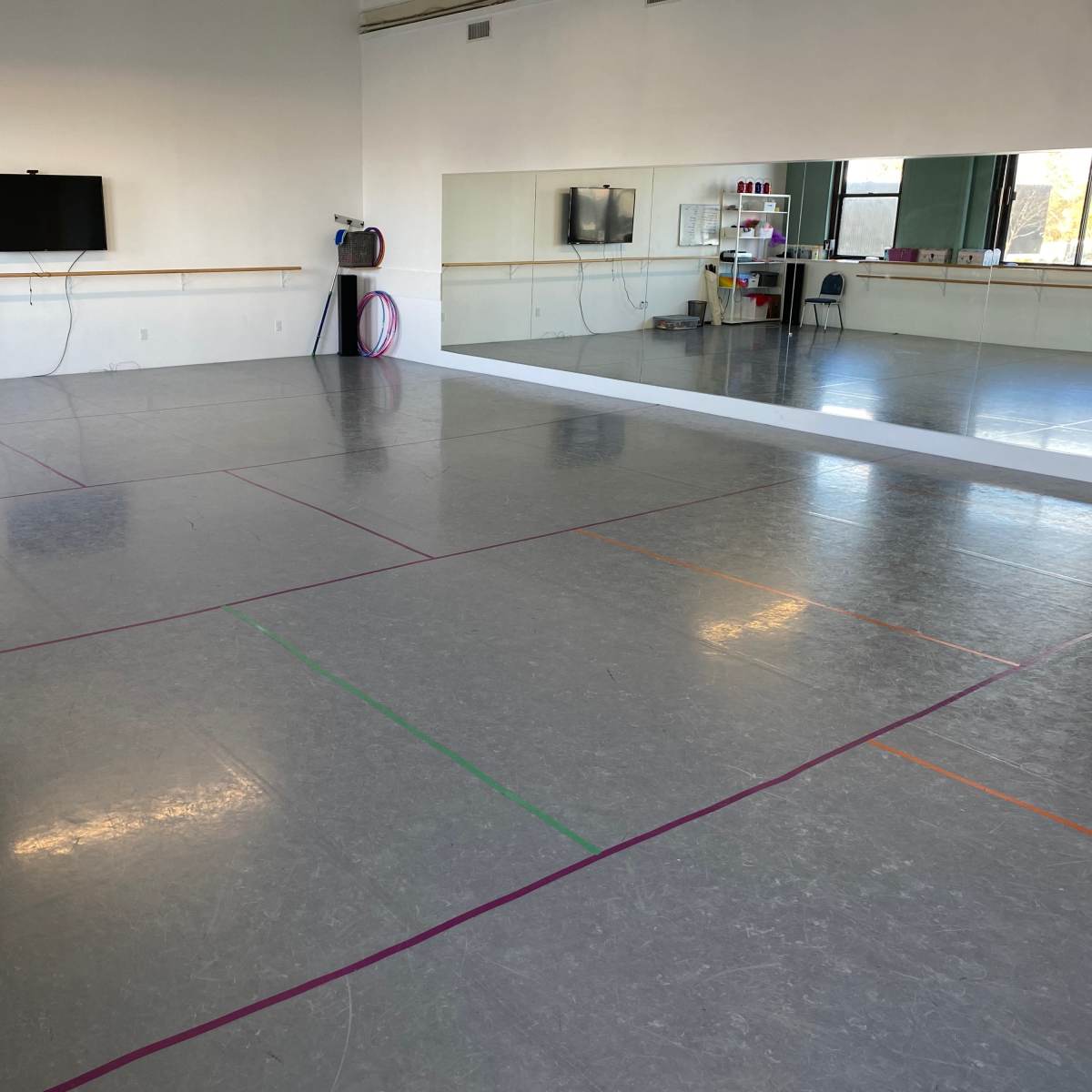 A spacious dance studio with a gray floor, wall mirrors, and minimal decor, featuring marked lines in various colors on the floor.