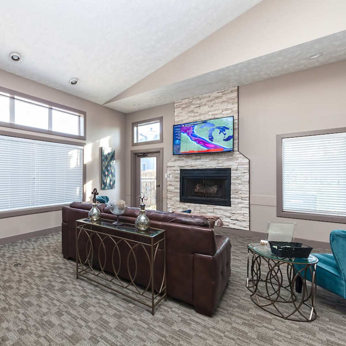 A modern living room with large windows, a stone fireplace, a television mounted on the wall, and a combination of seating and tables.