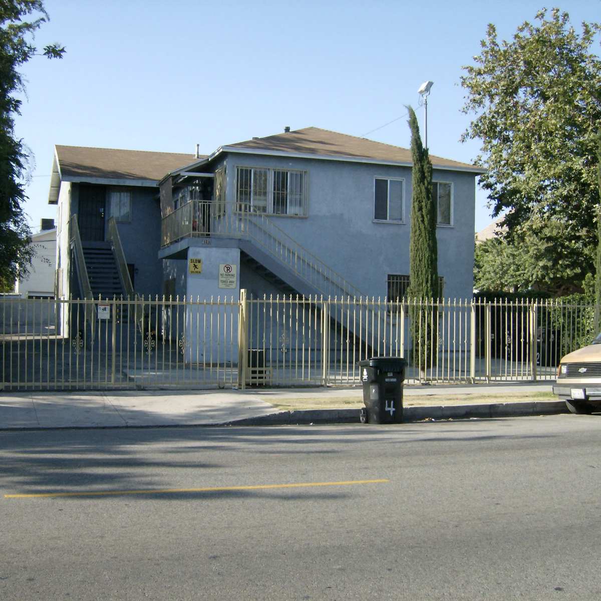 A two-story blue residential building with a fenced yard and a staircase on the side, located along a street.