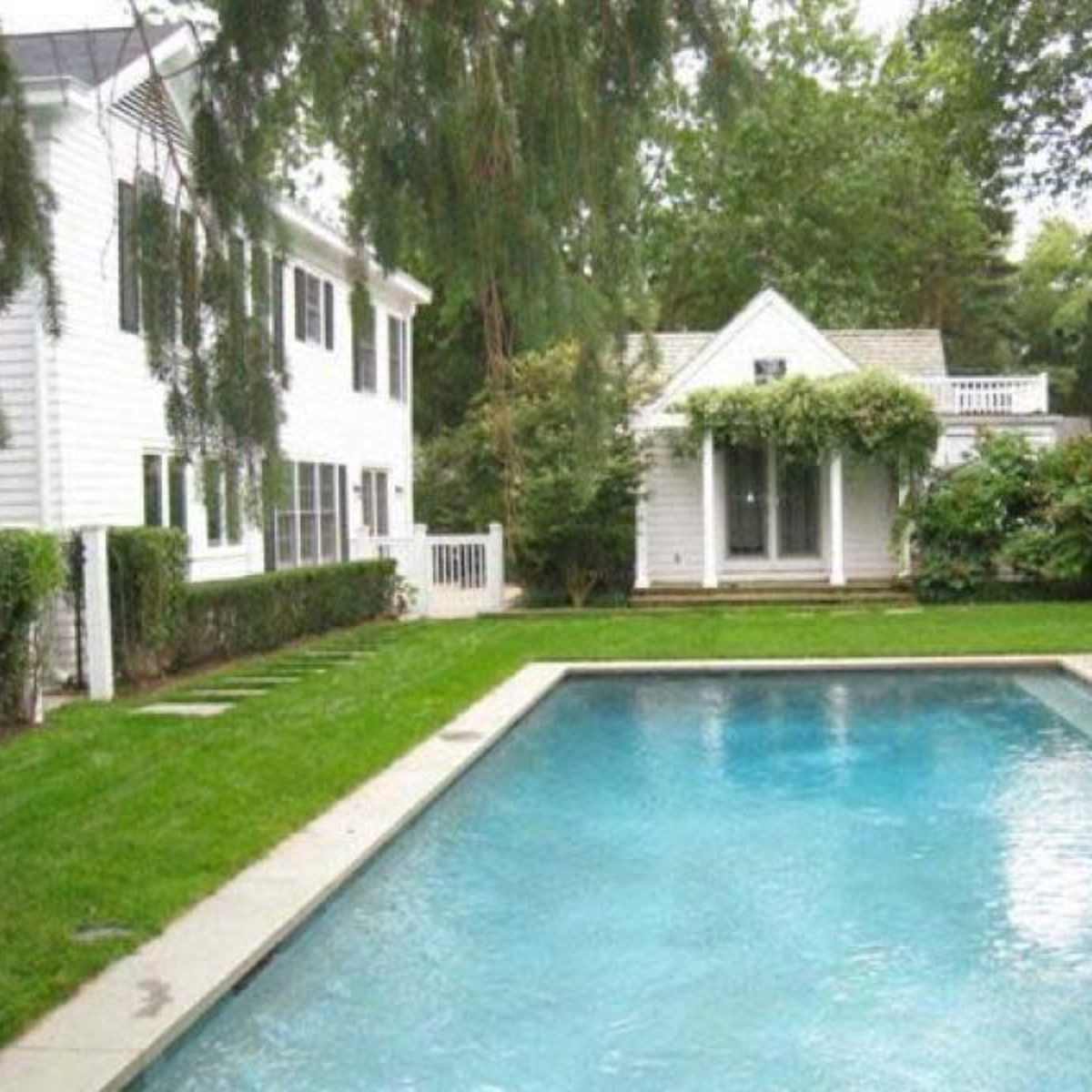 A well-maintained backyard features a clear swimming pool surrounded by greenery and a small white cottage in the background.