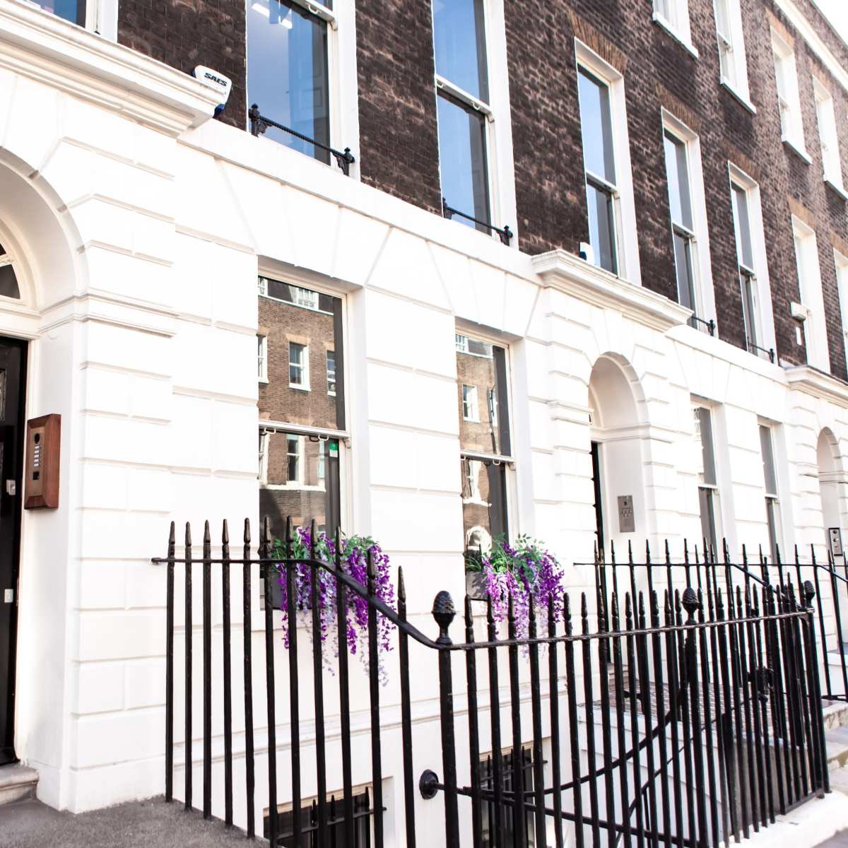 A row of Georgian-style townhouses with a black door and white exterior, featuring wrought iron railings and window boxes with purple flowers.