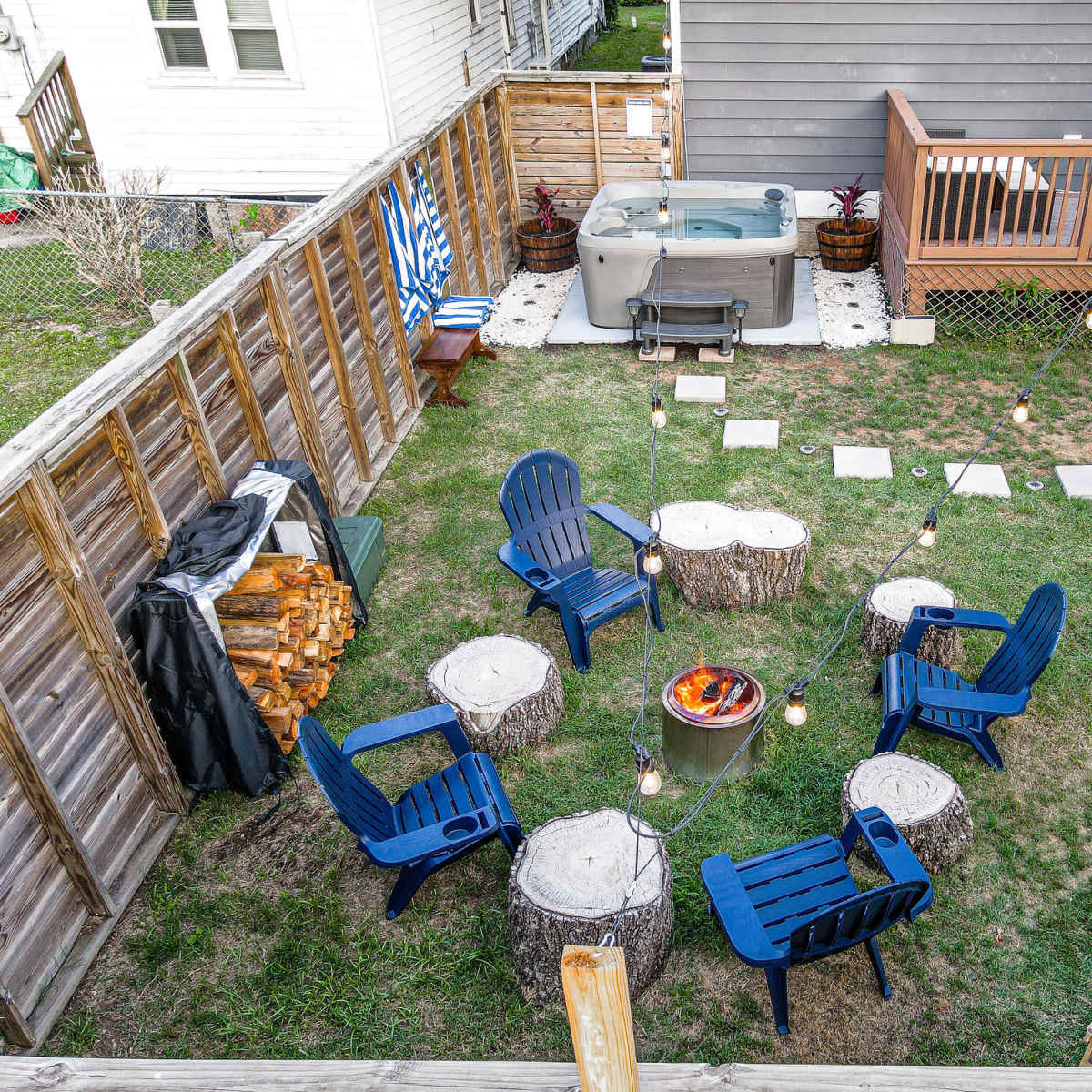 A backyard scene features circular seating made from tree stumps, a fire pit in the center, and a hot tub surrounded by decorative lights and wooden decking.